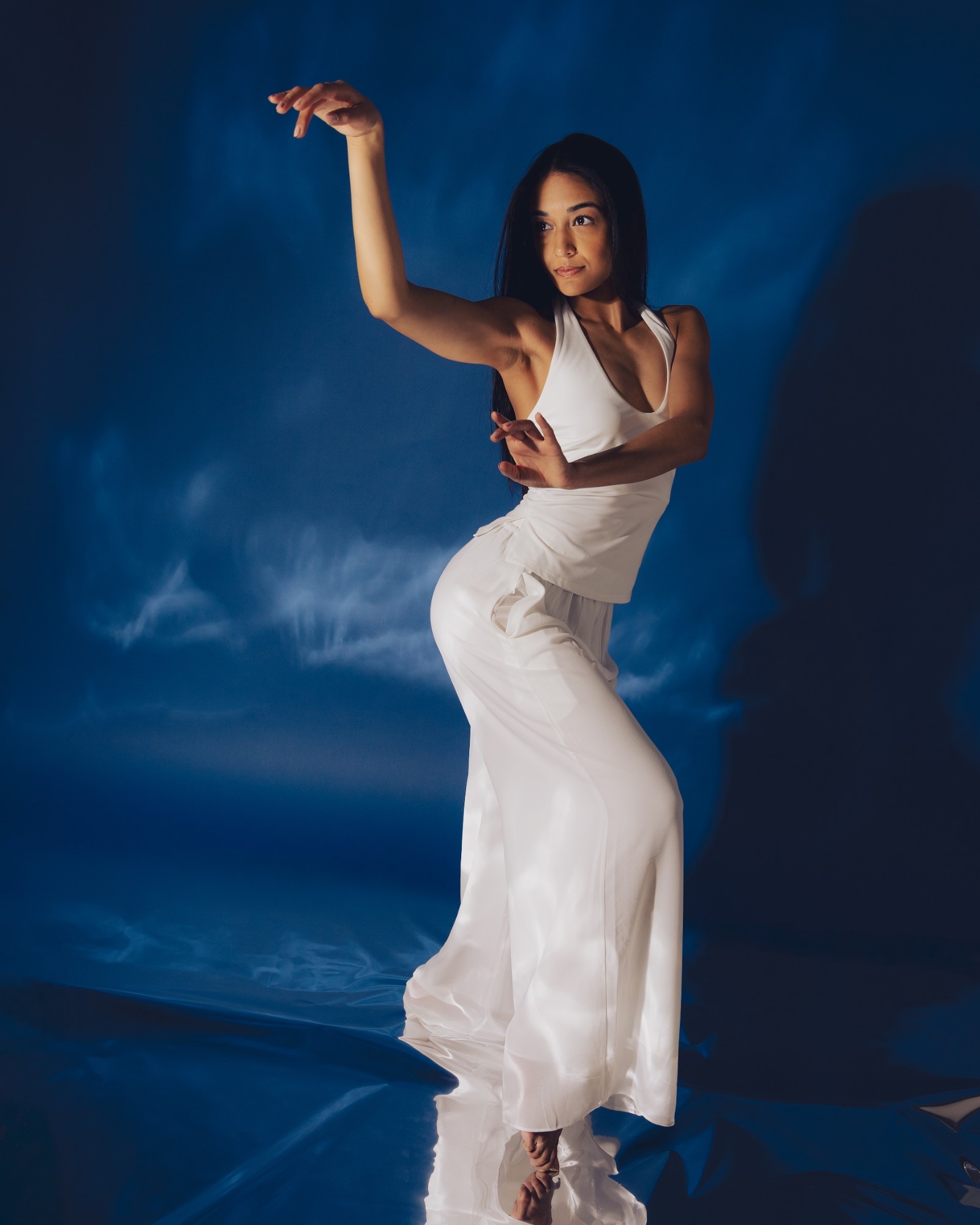 Shivani in flowing white, one arm raised, posed on a reflective floor against a deep blue backdrop